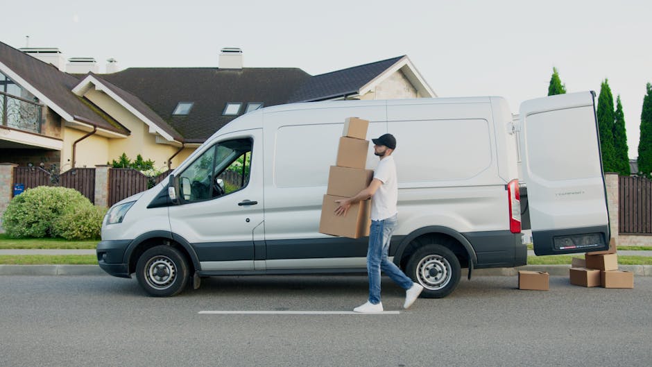 A man wearing casual clothing and a face mask is engaged in a home relocation process outside a residential property. He is carrying three stacked cardboard boxes towards a white panel van parked on the street in front of the house. The van, used for furniture transport and moving logistics, has its side door open, revealing the interior space suited for packing and moving items. Several additional cardboard boxes are placed on the paved road nearby, ready for loading. The residential area features modern houses, with a driveway, green shrubs, and tall trees in the background, illuminated by natural daylight. This scene depicts the loading process involved in house removals, with the man and Van Clapham facilitating relocation services efficiently and professionally.