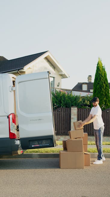 A man dressed in casual clothing is seen outside a residential property, preparing to load cardboard boxes into the rear compartment of a white commercial van parked on the street. The open van door reveals a spacious interior suitable for home relocation, with the man lifting a box towards the van. Several cardboard boxes of varying sizes are stacked on the pavement beside him, some sturdy and sealed with packing tape, indicating they are packed for moving. The surrounding environment includes a suburban house with a pitched roof, a wooden fence, and well-maintained greenery such as tall shrubs and trees. The scene is illuminated by natural daylight, demonstrating a typical moving process involving furniture transport and packing assistance provided by Man and Van Clapham, offering professional removals services in the area.