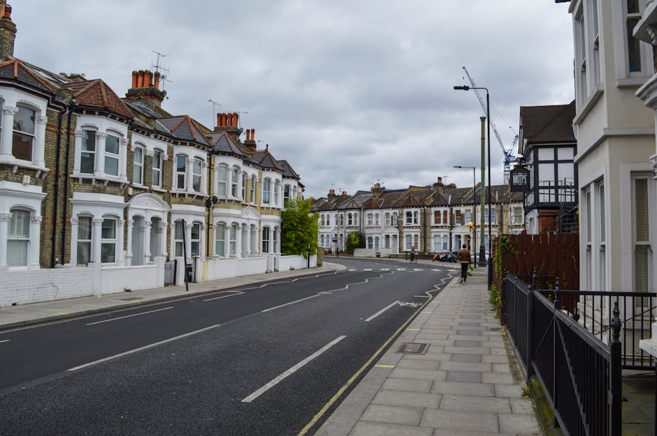 A quiet residential street in Clapham with a row of Victorian-style terraced houses featuring white facades, bay windows, and red tiled roofs. The pavement on the right side of the image runs parallel to the street, with a black metal fence bordering some of the properties and a few small plants visible. The road is marked with lane lines and has no parked vehicles on the near side, while a few cars are visible further down the street. Overhead, there are street lamps, and the sky is cloudy with grey clouds, indicating overcast weather. In this context of home relocation and furniture transport, [COMPANY_NAME] may assist with moving logistics, packing, and loading processes within this residential area, supporting house removals near Clapham Common SW4.
