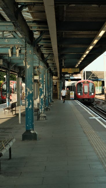 A railway station platform with a red passenger train on the right side, positioned parallel to the platform. The platform features a grey, textured walking surface with tactile paving for visually im